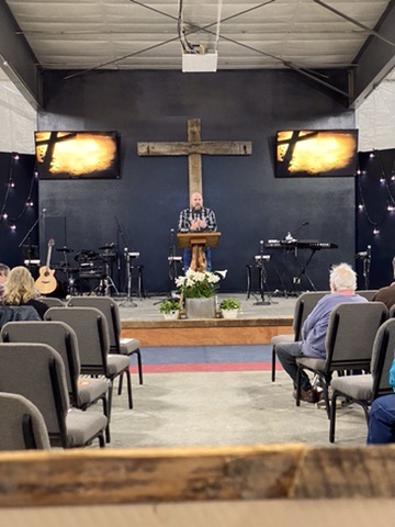 Pastor Toby Goodman preaching from the pulpit beneath a wooden cross at Gallatin River Fellowship