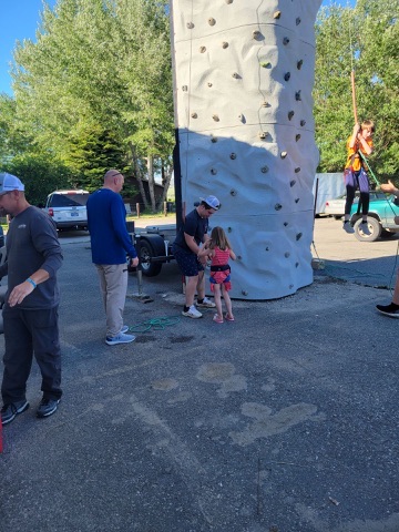 A father helping his young daughter try a portable climbing wall at a Gallatin River Fellowship community outreach
